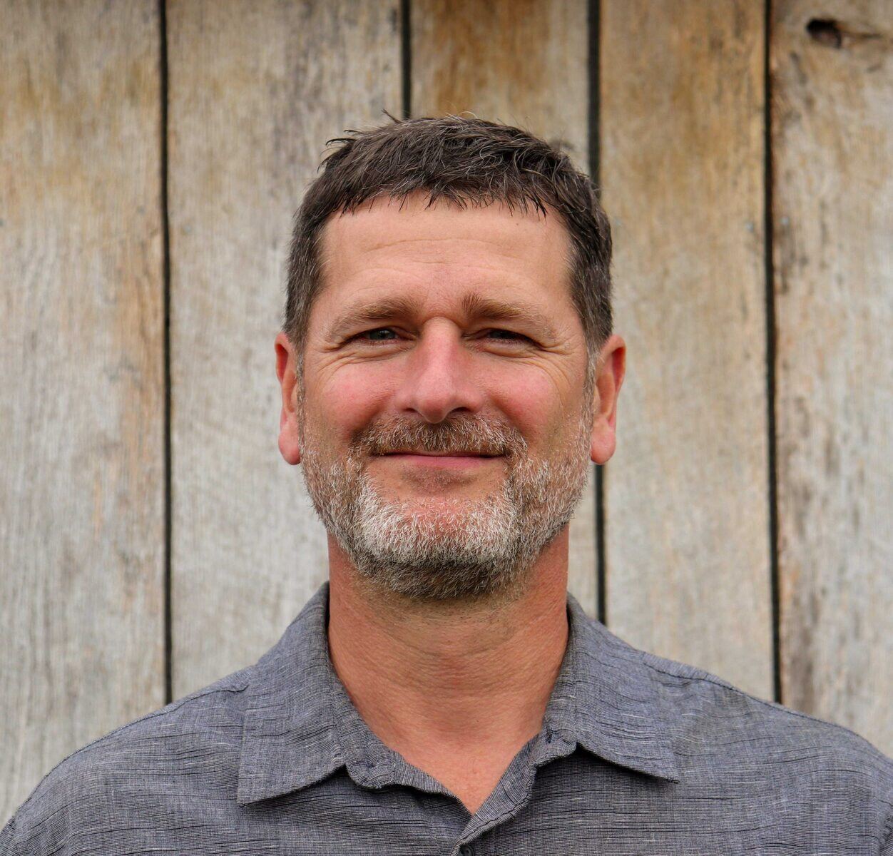 Outdoor portrait of a smiling man with a wooden backdrop, part of the R Value Homes team in West Michigan